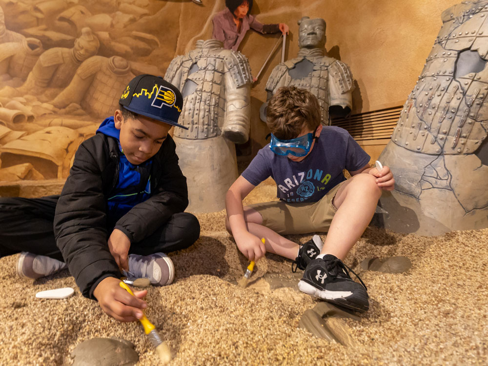 Children sitting in pretend dirt using brushes to wipe away dust. Replicas of Terra Cotta Warriors are in the background.